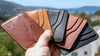 Bir elin fan şeklinde tuttuğu çeşitli Pueblo Kiremit, Natural, Siyah ve Buttero Kiremit, Siyah MZ Leather Craft minimalist deri kartlıklar. Bodrum manzarası eşliğinde el dikimi ve logolu kartlık çeşitlerinin görüntüsü.