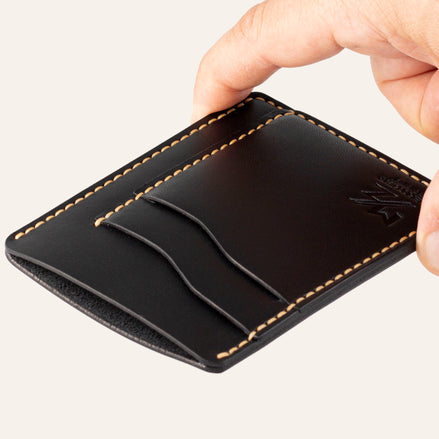 MZ Leather Craft dikey minimalist siyah Buttero deri kartlık, el ile tutulan hafif açılı boş ön görünüm, kıvrımlı kart yuvaları ve sarı saraç dikişi detayları | The Vertical Minimalist Cardholder - Black Buttero Leather by MZ Leather Craft, empty front view held by hand at a slight angle, showcasing curved card slots and yellow saddle-stitching details