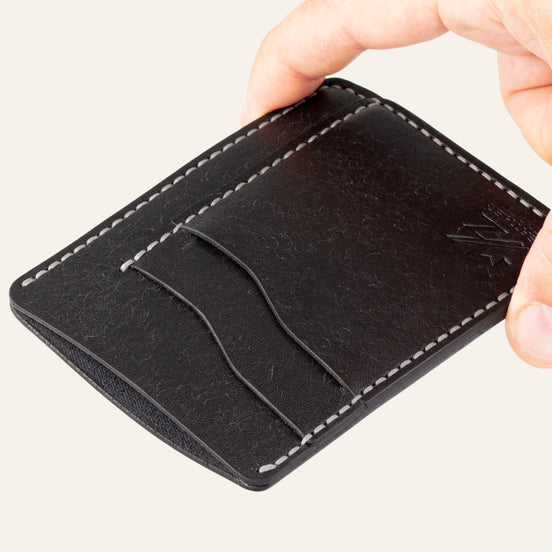 MZ Leather Craft dikey minimalist siyah Pueblo deri kartlık, el ile tutulan hafif açılı boş ön görünüm, kıvrımlı kart yuvaları ve gri saraç dikişi detayları | The Vertical Minimalist Cardholder - Black Pueblo Leather by MZ Leather Craft, empty front view held by hand at a slight angle, showcasing curved card slots and gray saddle-stitching details